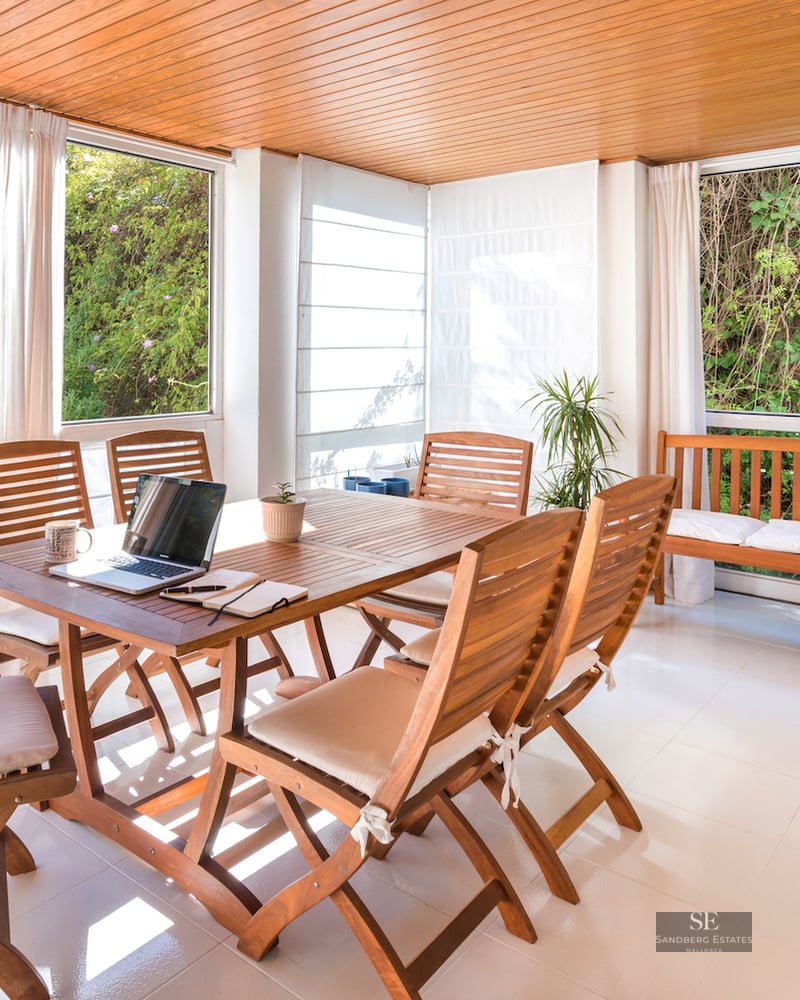 A sunlit terrace room with a wooden dining table, chairs, and bench, overlooking lush green garden foliage.
