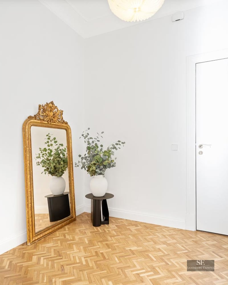 Bright foyer with white walls, herringbone wood floors, a large gold mirror, and a white door.