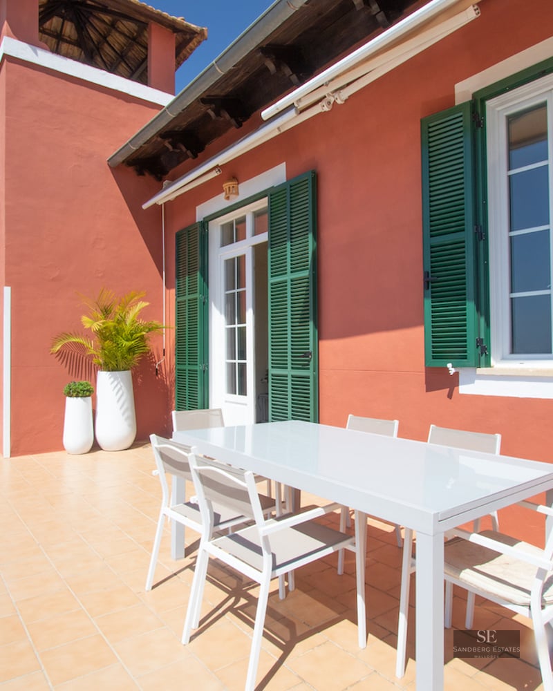 A sunlit terrace with terracotta walls, green shutters, and a white modern dining set under a clear blue sky.