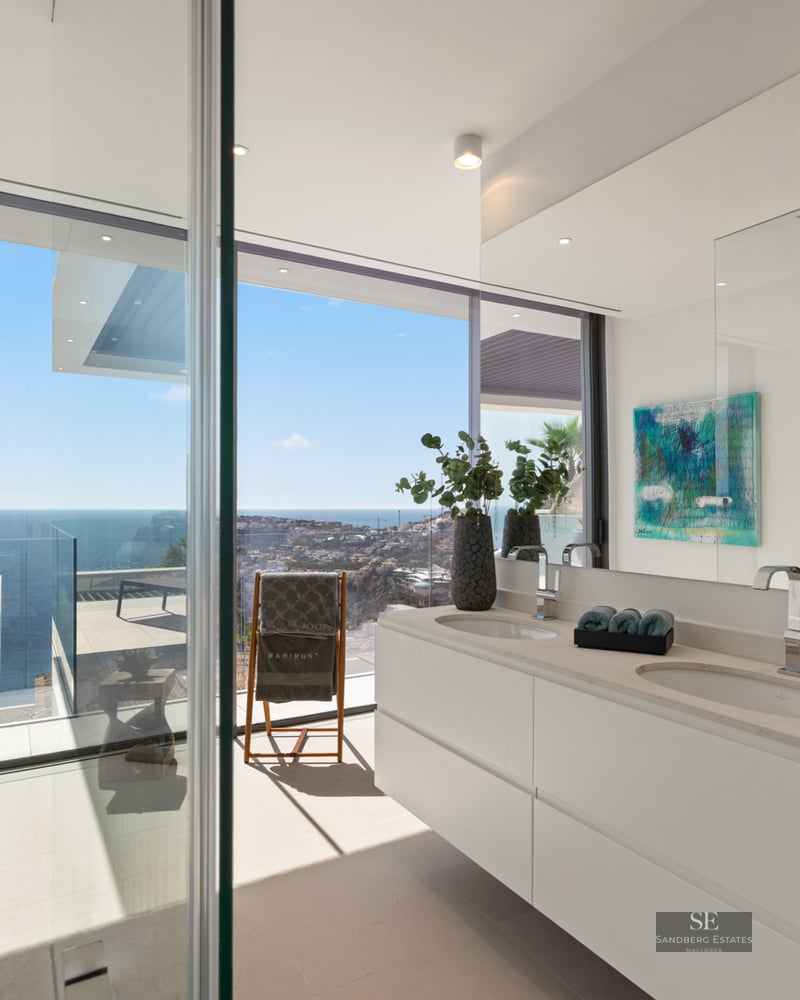 Modern bathroom featuring a double white vanity and floor-to-ceiling windows overlooking the Mediterranean sea and coastline.