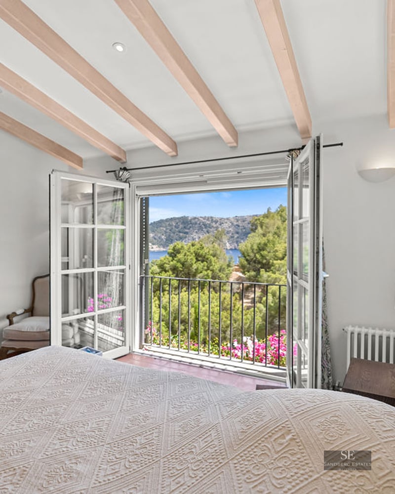 Bright bedroom with wooden ceiling beams and open balcony doors revealing a lush green hillside and sea view.