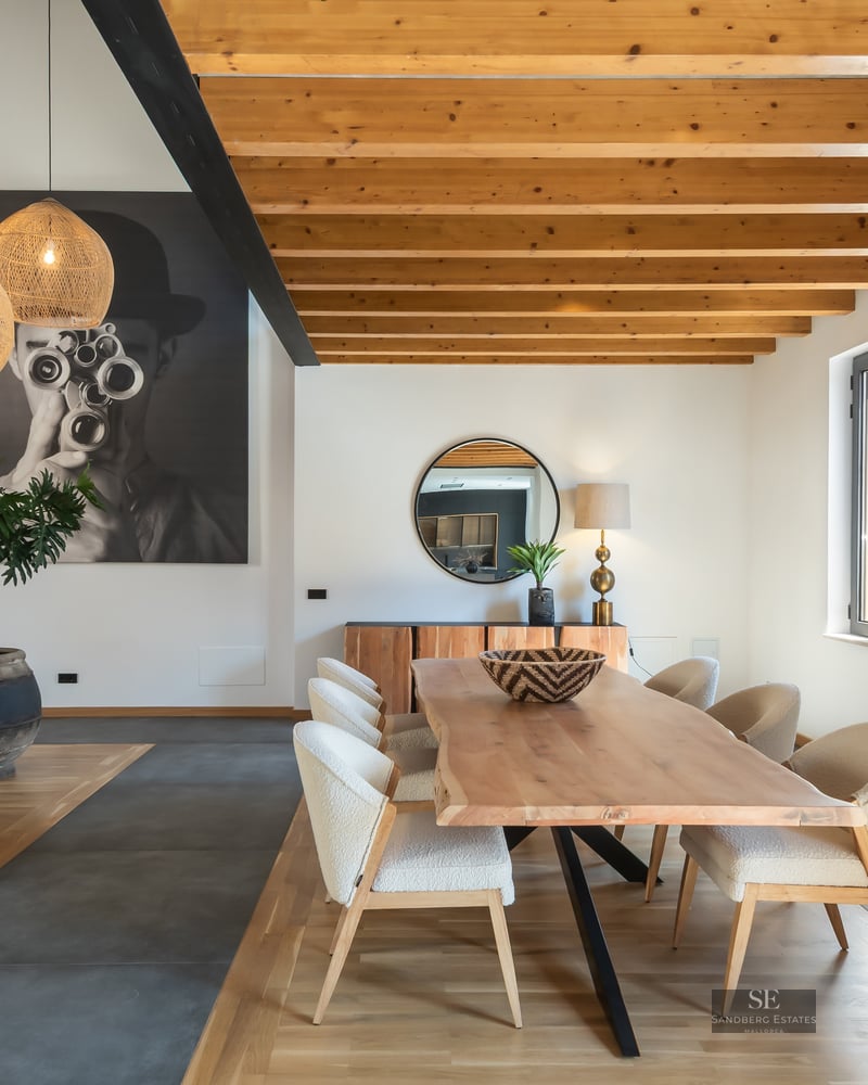 Elegant dining room featuring a live-edge wooden table, upholstered chairs, exposed ceiling beams, and large wall art.