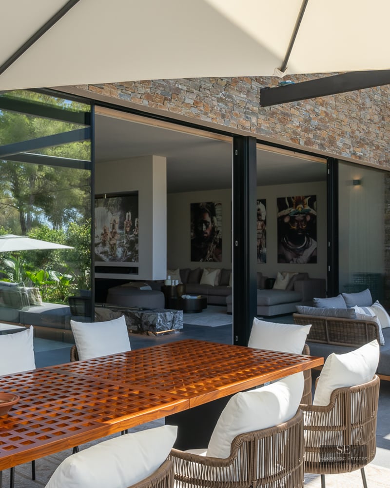 Modern terrace featuring a large wooden dining table, grey sofas, and floor-to-ceiling glass doors to the living room.