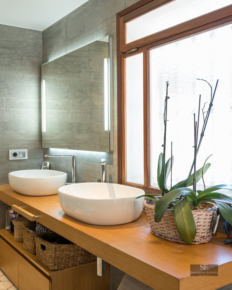 Contemporary bathroom with two white vessel sinks on a wooden vanity, grey tiled walls, and a large backlit mirror.