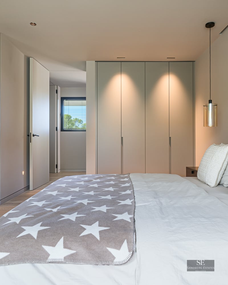 Bright modern bedroom featuring a large bed with white linens, grey star throw, and built-in closets with warm lighting.