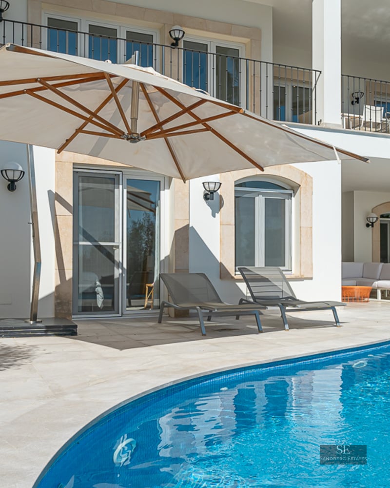 Piscine bleue incurvée avec terrasse en pierre blanche, grand parasol beige et transats devant une villa blanche.