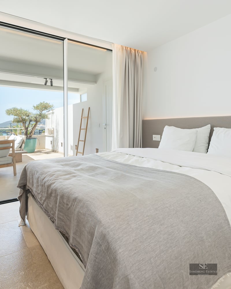 Bright modern bedroom with a large bed, stone floors, and floor-to-ceiling glass doors opening to a sunny terrace.