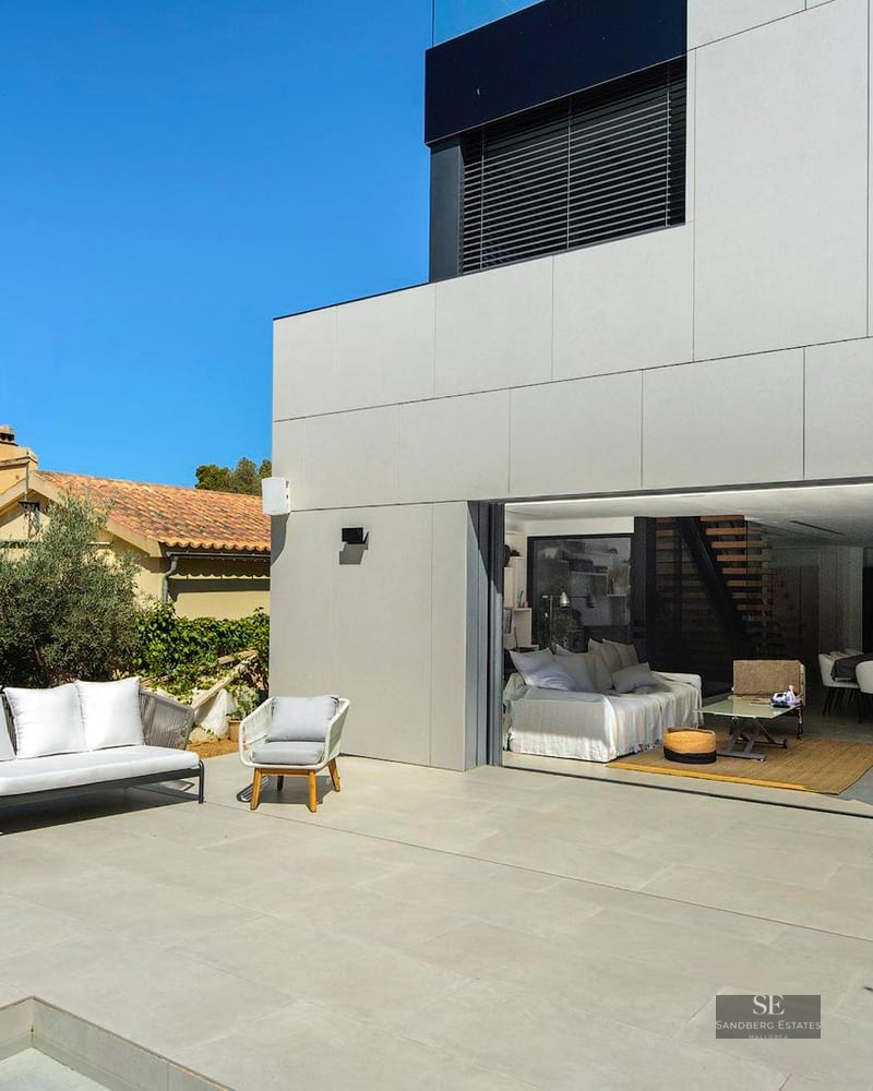 Modern outdoor terrace with white furniture next to a villa's open-plan living area and a stone wall.