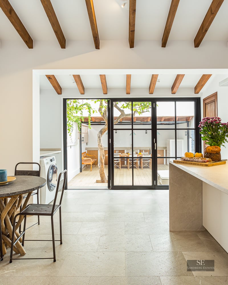 Bright kitchen with wood ceiling beams, stone island, and glass doors opening to a sunny outdoor patio with a tree.