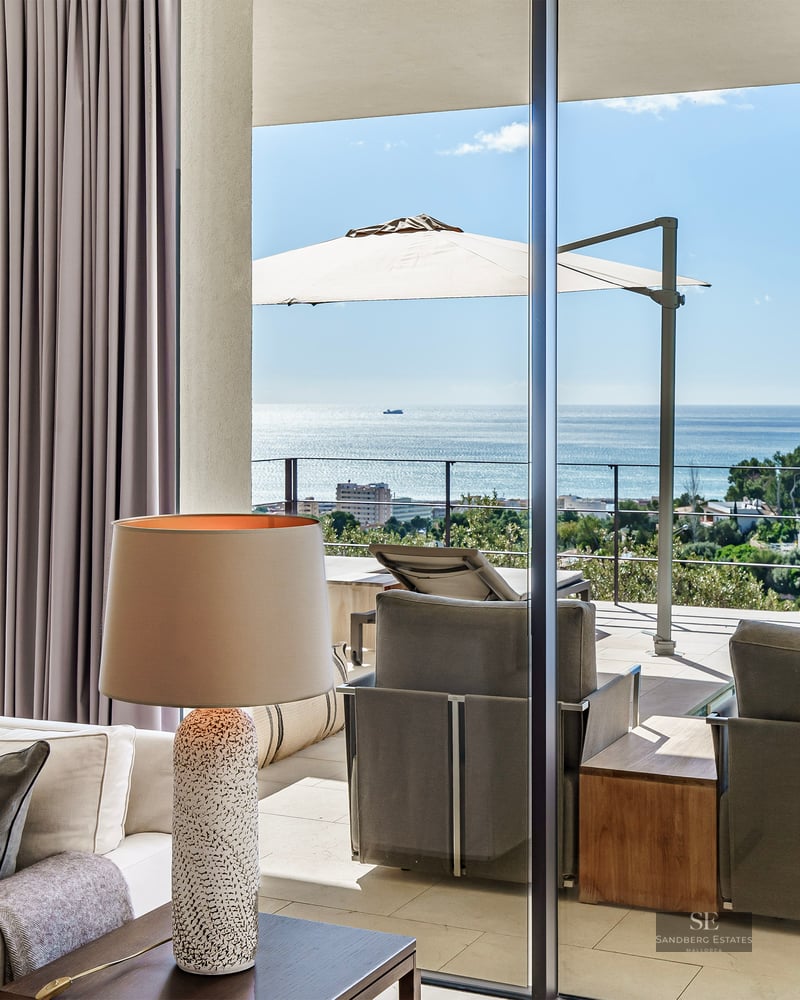 Elegant living room with beige sofas and floor-to-ceiling glass doors opening to a sunlit terrace with blue sea views.