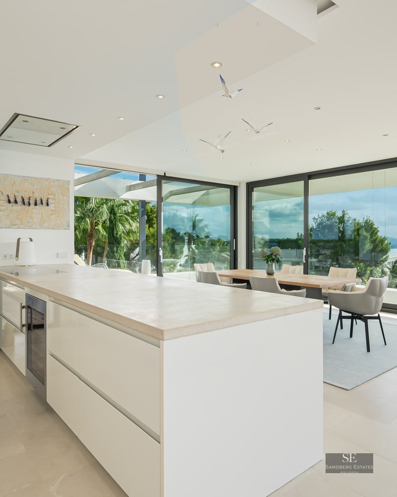 Bright modern white kitchen with large island, dining area, and floor-to-ceiling glass walls overlooking the ocean.