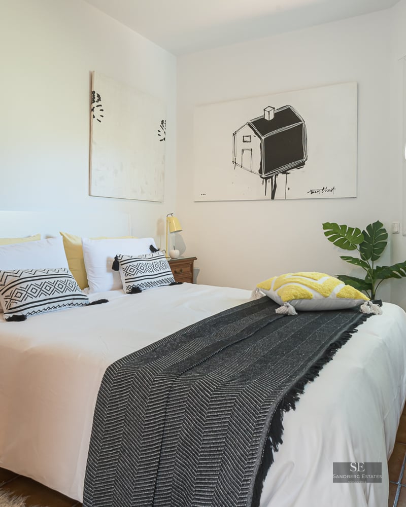 Modern bedroom with a white bed, patterned pillows, wooden nightstands, and tile flooring under natural light.