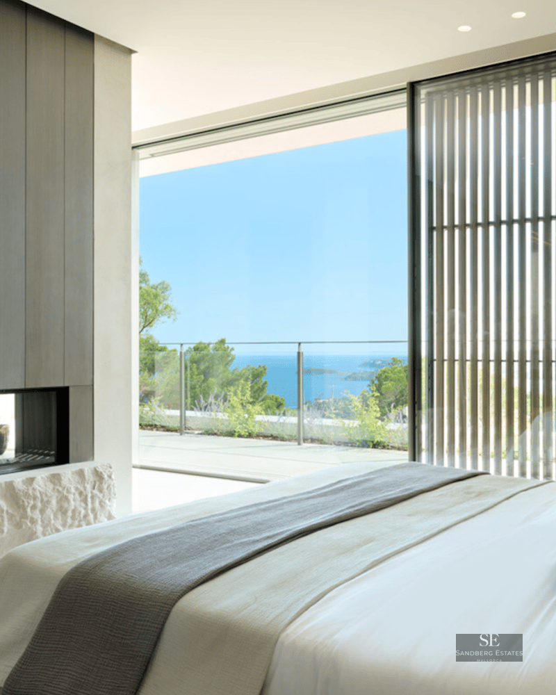 Luxury bedroom featuring floor-to-ceiling windows overlooking the ocean and a modern fireplace with a rough stone base.
