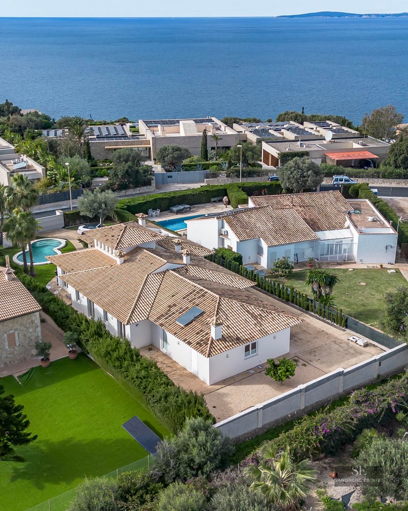 High-angle drone shot of a white villa with terracotta roof, green lawn, and the blue ocean in the background.