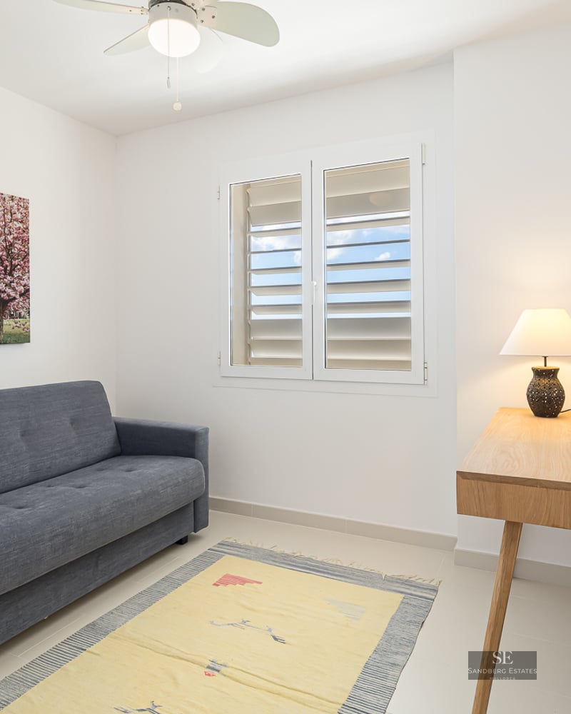 A minimalist room with a grey sofa, wooden desk with a lamp, a blue chair, and a window with white shutters.