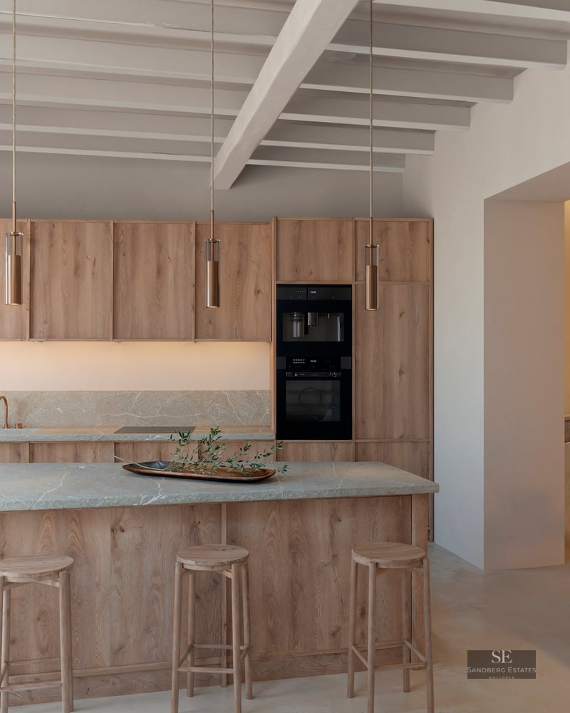 Luxury kitchen featuring light wood cabinets, stone island with stools, and exposed ceiling beams.