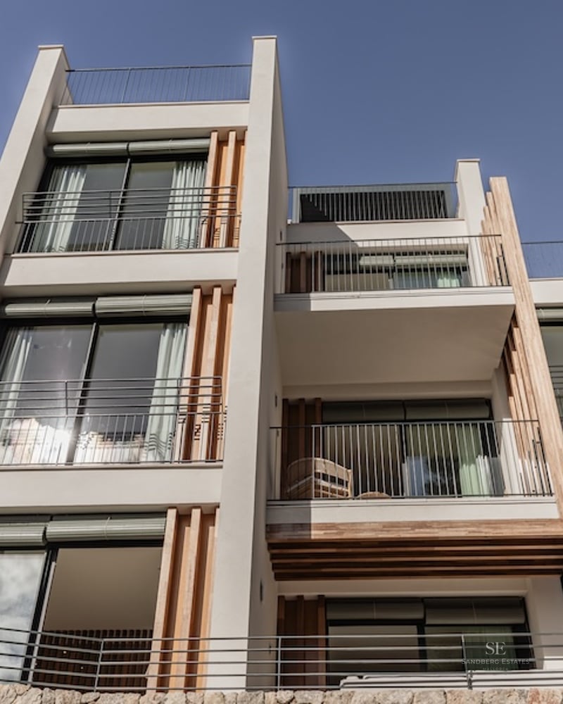Low-angle view of a white modern villa with wooden slats, large glass windows, and black metal balconies against a blue sky.