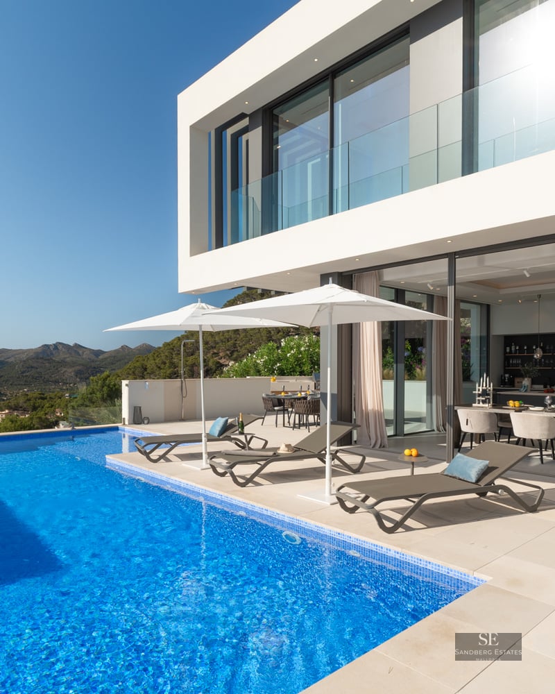 Luxury infinity pool and white modern villa with mountain views and designer loungers under white umbrellas.