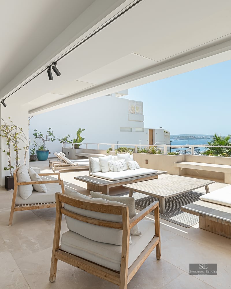 Spacious white terrace with wooden lounge furniture overlooking a marina and the blue Mediterranean sea.