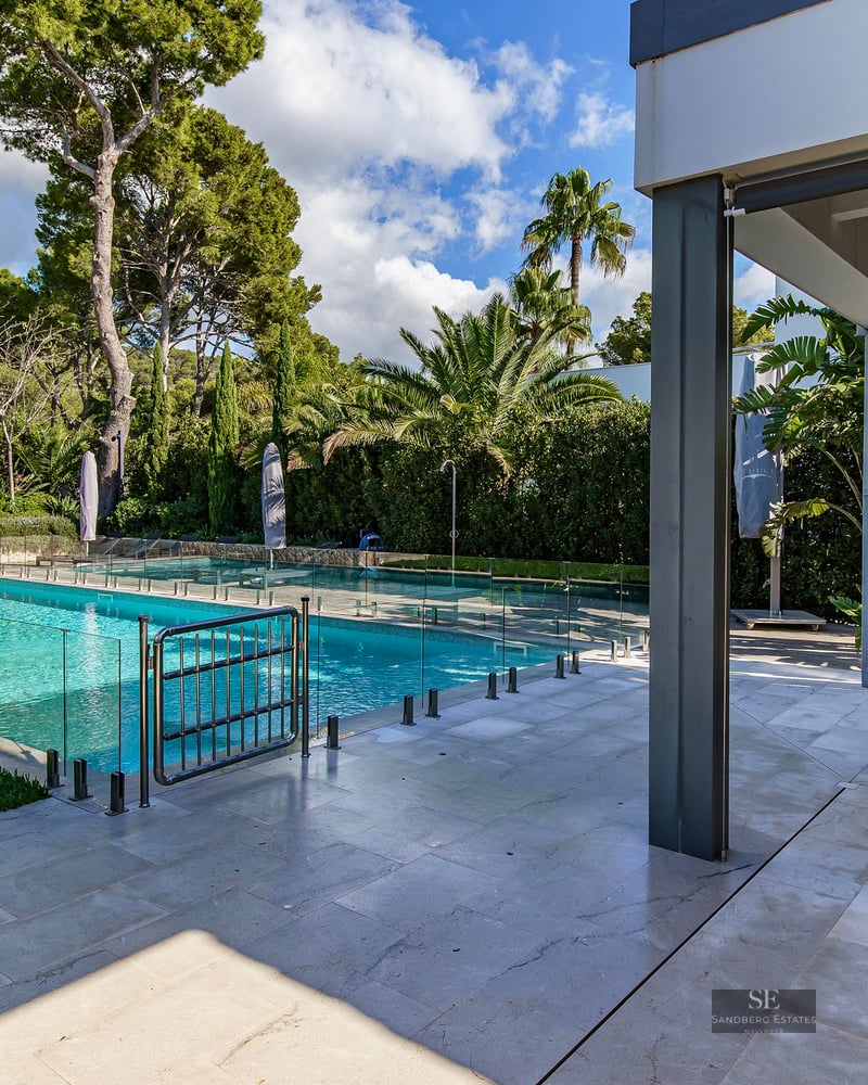 Piscine turquoise avec clôture en verre à côté d'une terrasse en pierre ombragée avec table et jardin tropical.