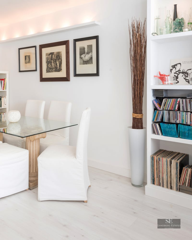 A bright dining room with a glass table, white chairs, and extensive white bookshelves filled with books and music.