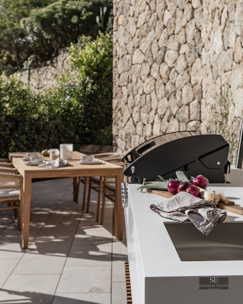 Modern outdoor kitchen with white countertop, sink, and grill next to a stone wall and wooden dining set.