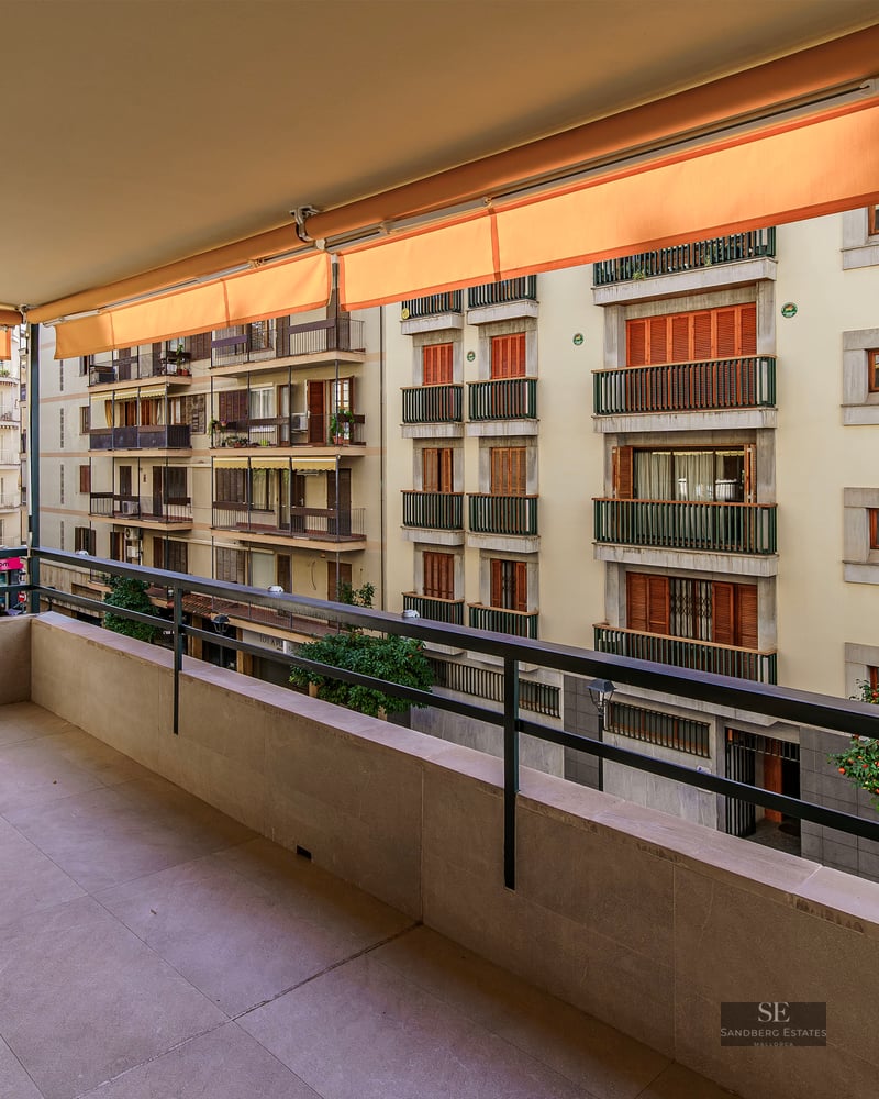 Large terrasse en pierre avec garde-corps métallique et stores orange donnant sur des immeubles résidentiels.