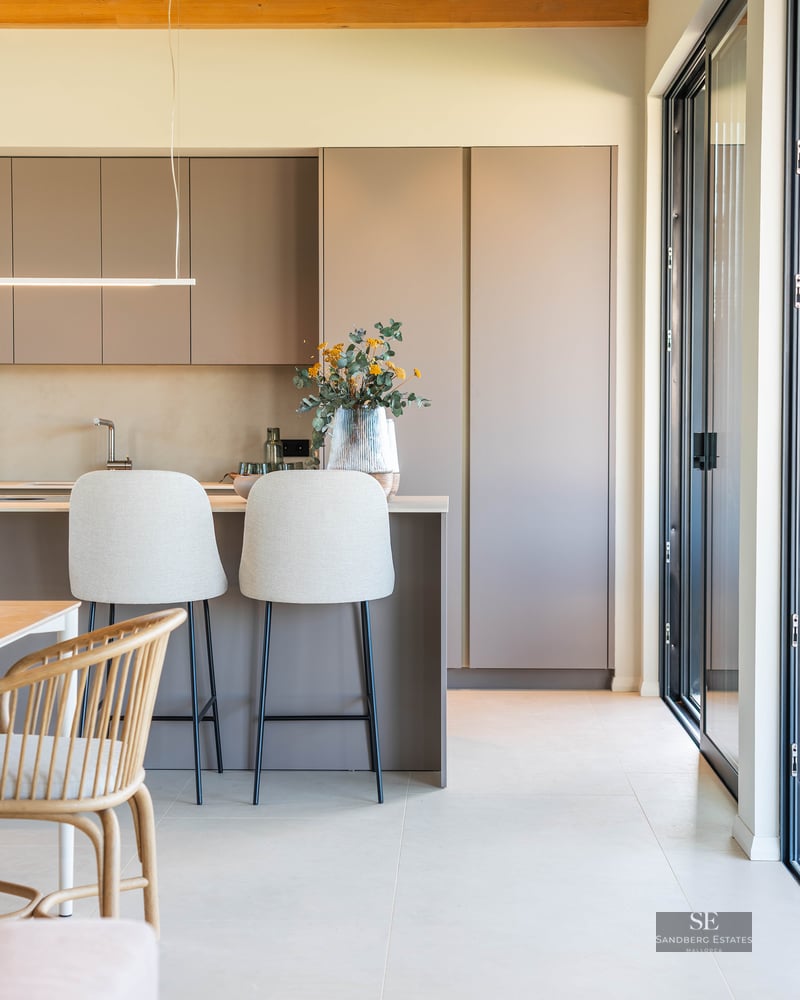 A modern kitchen featuring matte taupe cabinets, an island with two light-colored bar stools, and a wooden ceiling beam.