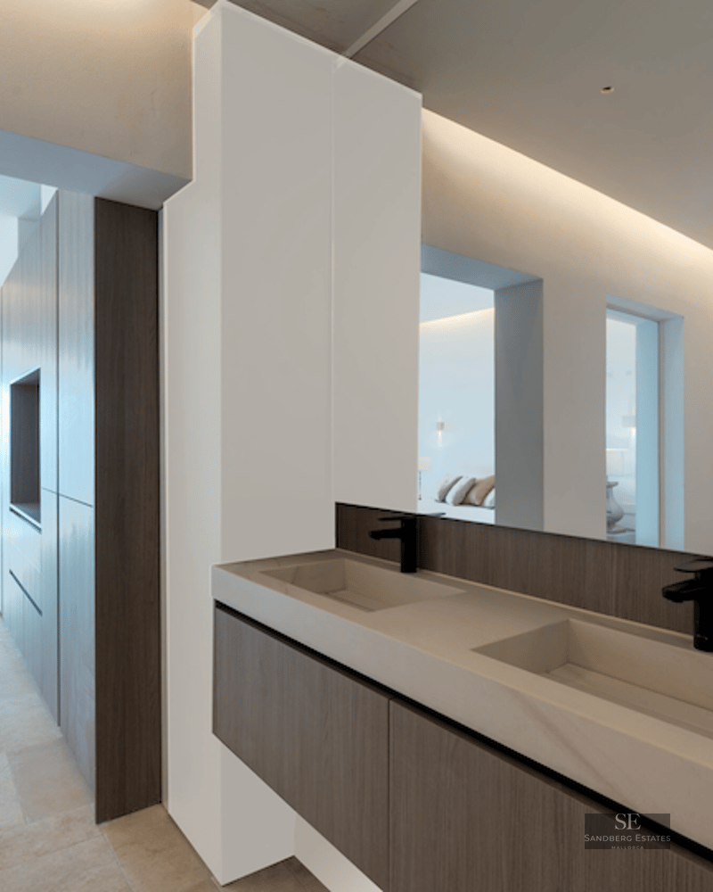 Modern master bathroom featuring double stone sinks, dark wood vanity, and a view through to a bedroom and the sea.