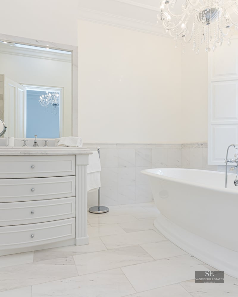 Luxury white marble bathroom featuring a freestanding tub, crystal chandelier, and large window with natural light.