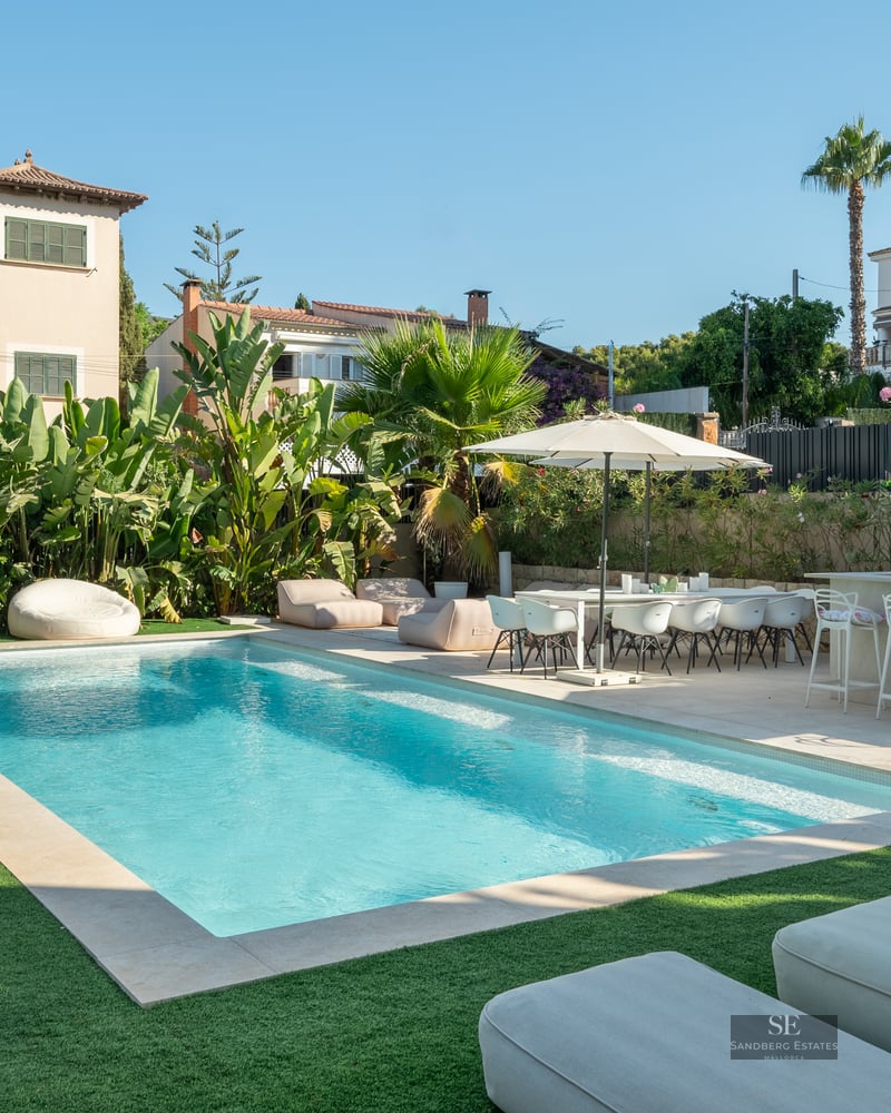 A sparkling rectangular swimming pool surrounded by a green lawn, outdoor dining set, bar, and lush tropical plants.