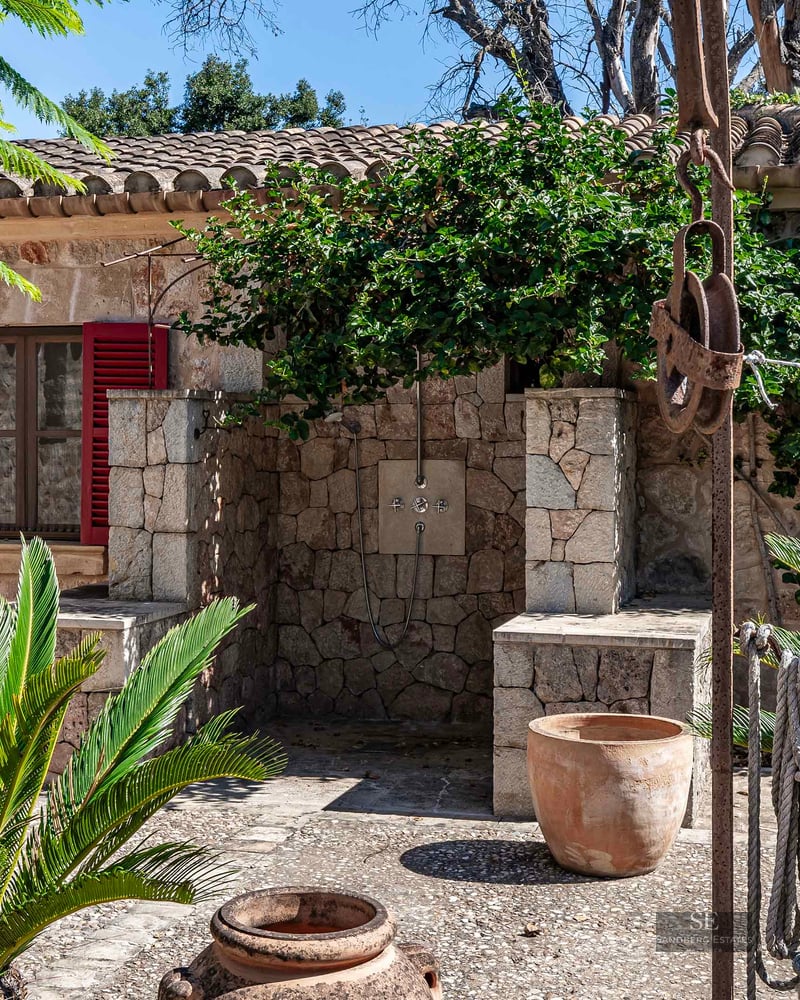 Outdoor stone shower area with red window shutters, terracotta pots, and lush green plants in a Mediterranean courtyard.