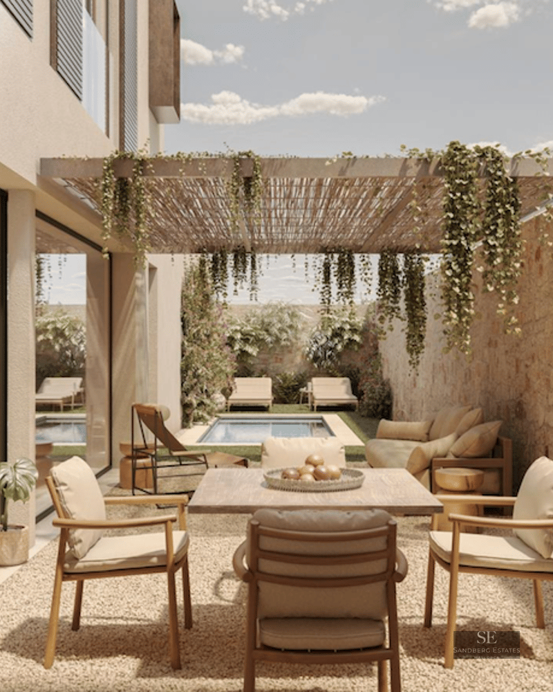 Modern outdoor terrace with a wooden dining table, a stone wall, a pergola with plants, and a pool in the background.