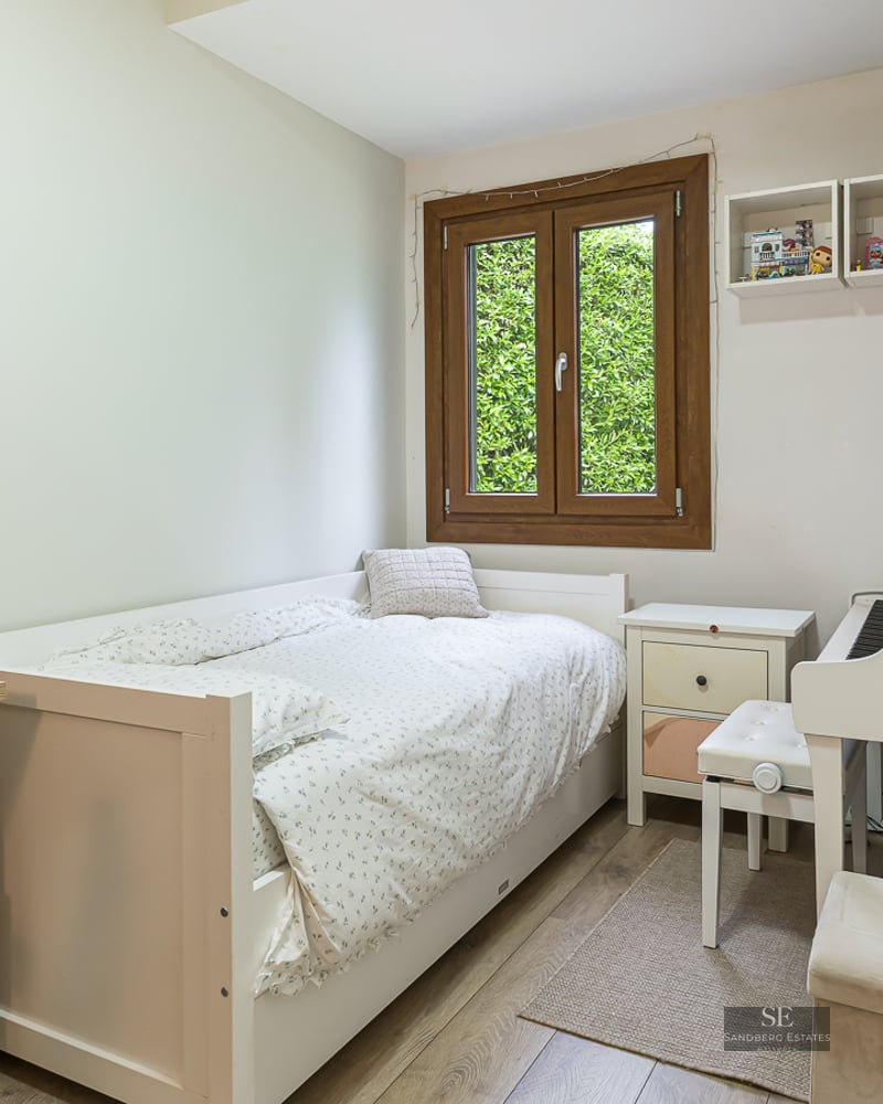 Bright bedroom with a single white bed, a desk, a white digital piano, and a window showing green foliage.
