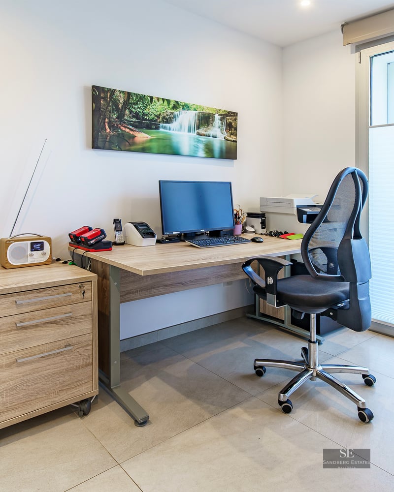 Home office featuring a light wood desk, ergonomic chair, computer setup, and large window with blinds.