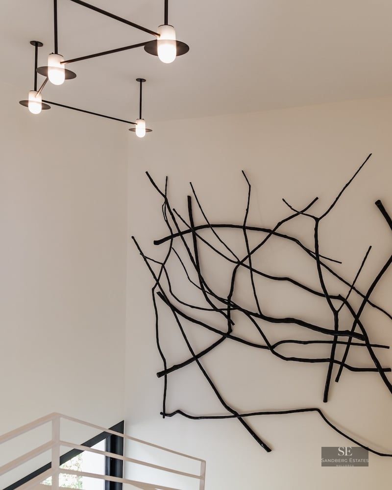 Black branch-like wall sculpture next to a modern geometric chandelier in a white minimalist room with stairs.