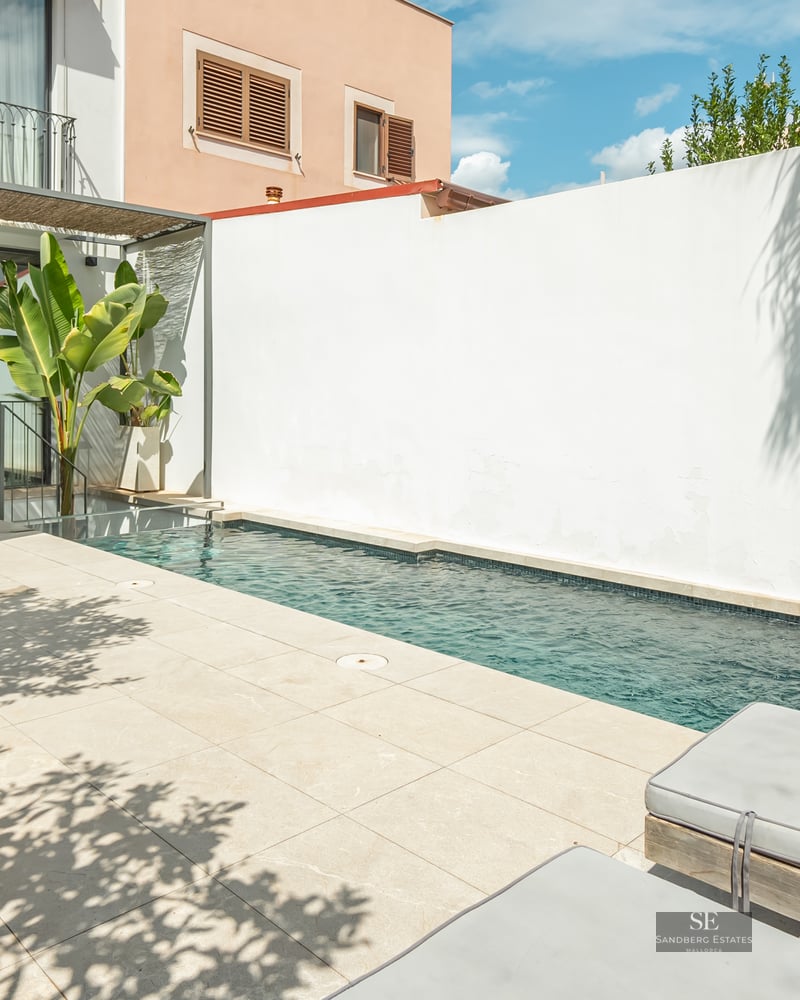 A narrow blue swimming pool on a light-colored tiled terrace with sun loungers and an outdoor dining area.