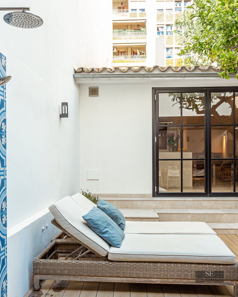 An outdoor shower with patterned blue tiles next to sun loungers on a wooden deck with a lemon tree.