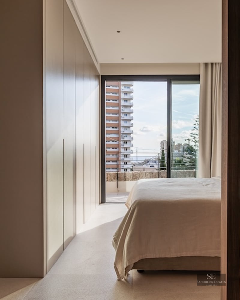 Modern bedroom with large built-in wardrobes, a bed with light linens, and a balcony window showing city buildings.