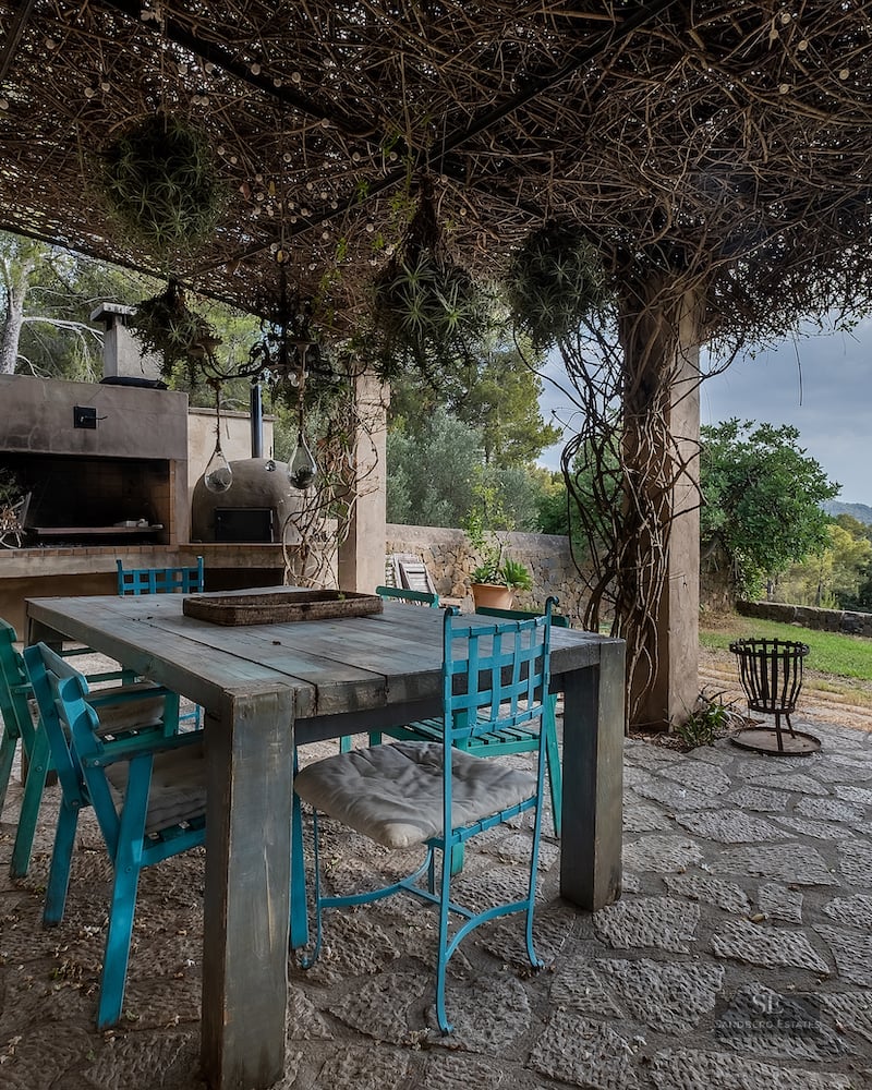 Rustic stone terrace with turquoise chairs, wooden table, and BBQ under a vine-covered pergola overlooking hills.