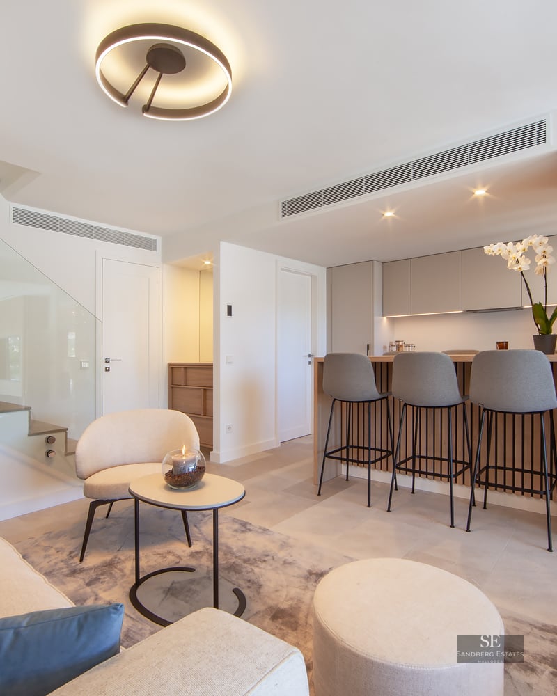 Modern living room with cream sofa, glass staircase, and a wood-paneled breakfast bar with grey stools.