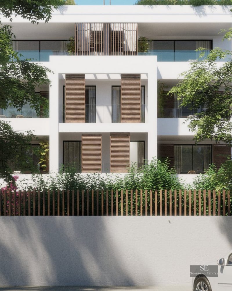 White four-story modern building with wooden shutters and glass balconies framed by large green trees.
