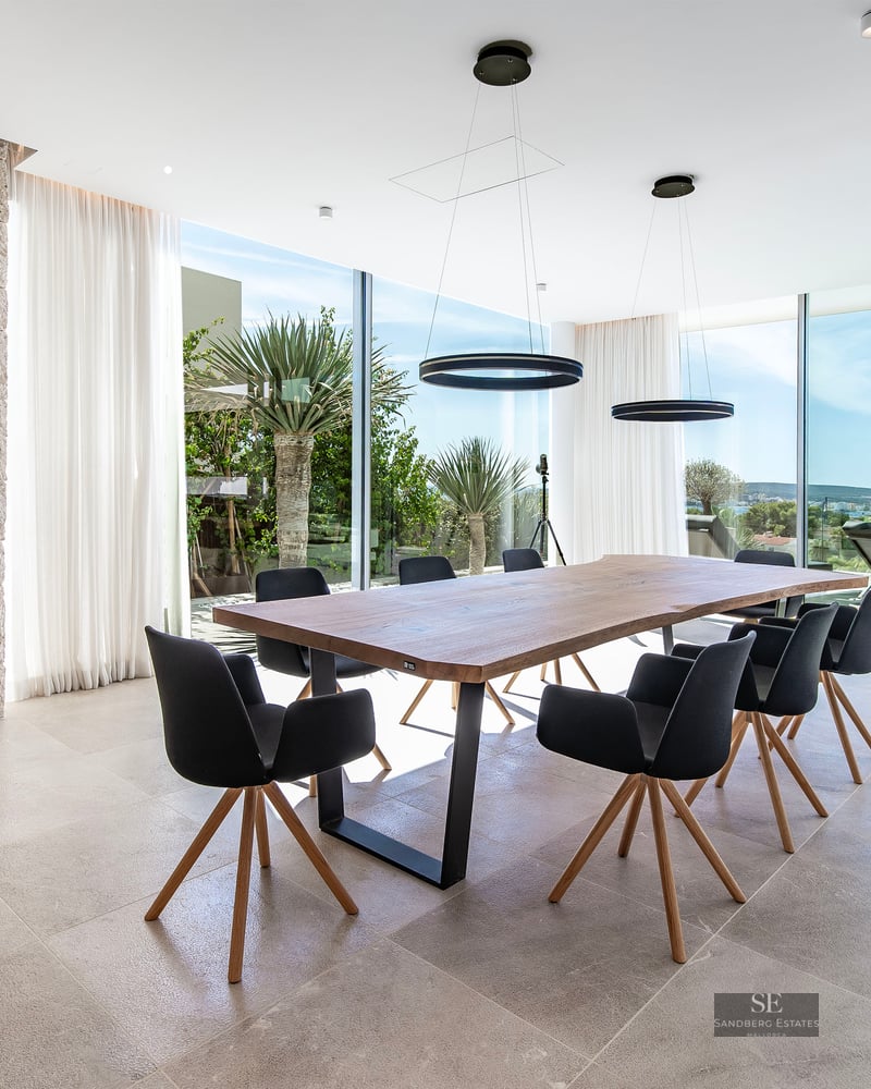 A modern dining room featuring a large wooden table, black chairs, a stone wall with a skull sculpture, and large windows.