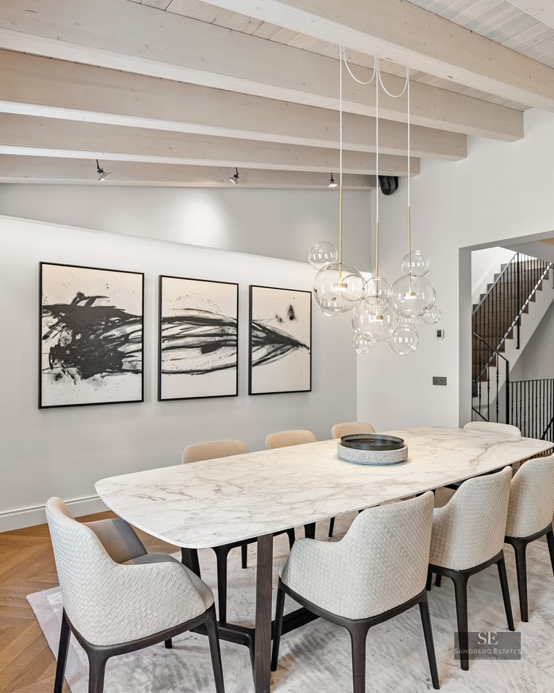 Modern dining room featuring a white marble table, designer chairs, bubble chandelier, and exposed wood ceiling beams.