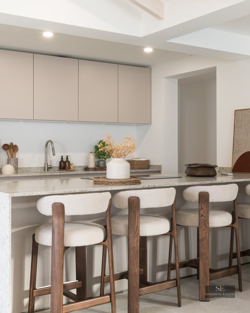 A contemporary kitchen featuring beige cabinetry, a white stone island, and four wood and fabric bar stools.