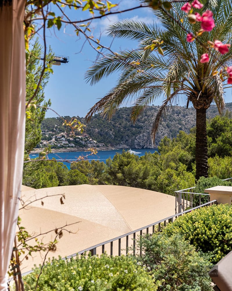 A luxury balcony terrace with lounge chairs and dining area overlooking a blue sea bay and green mountains.