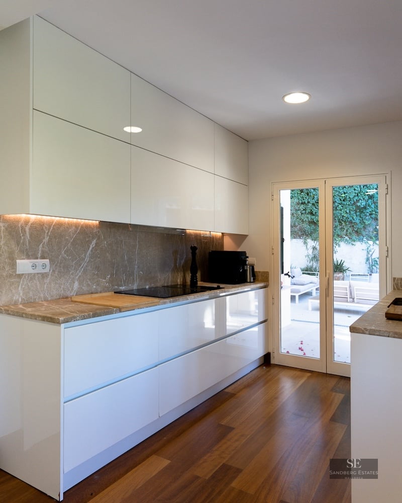 Modern white kitchen featuring marble countertops, wood flooring, and glass doors leading to an outdoor terrace.