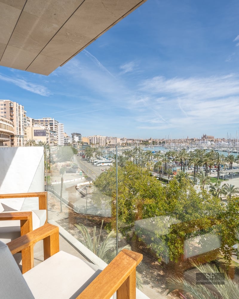 Modern balcony with wooden armchairs and glass railing overlooking a panoramic marina with yachts and city buildings.