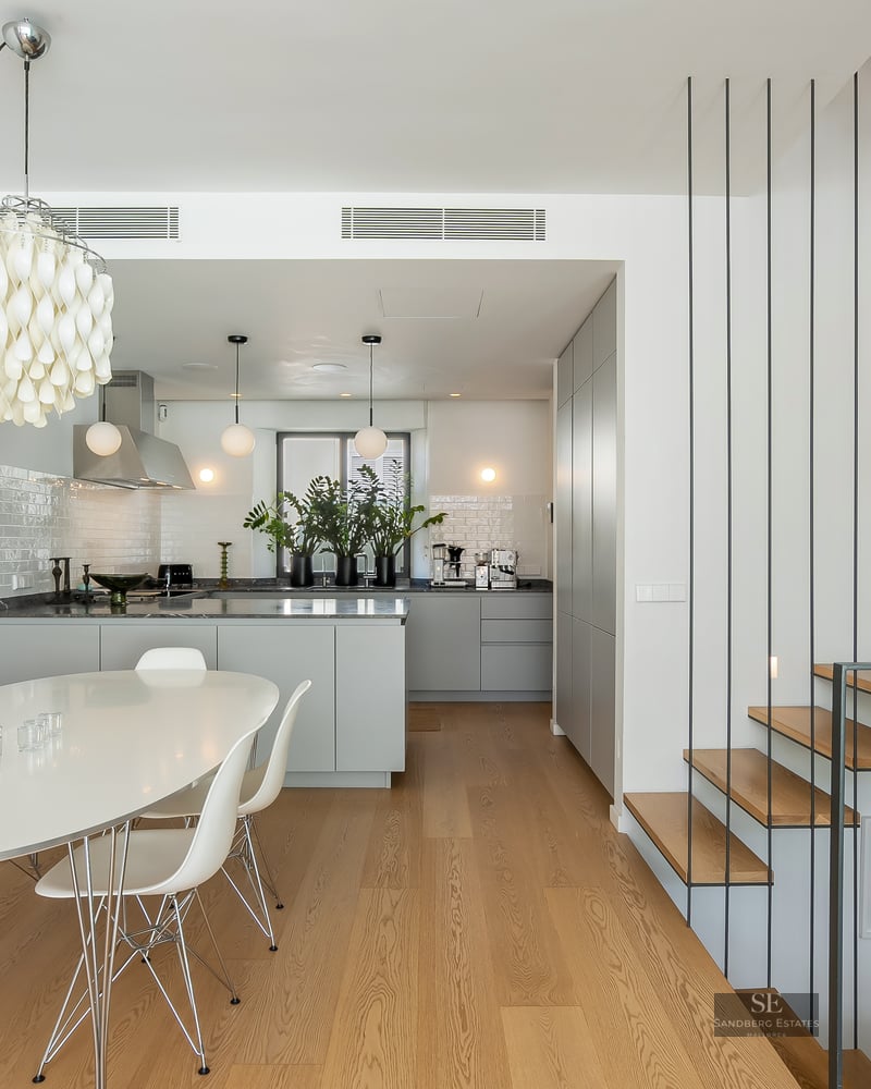 White dining room with oval table, Eames chairs, designer chandelier, grey kitchen, and wood/metal staircase.
