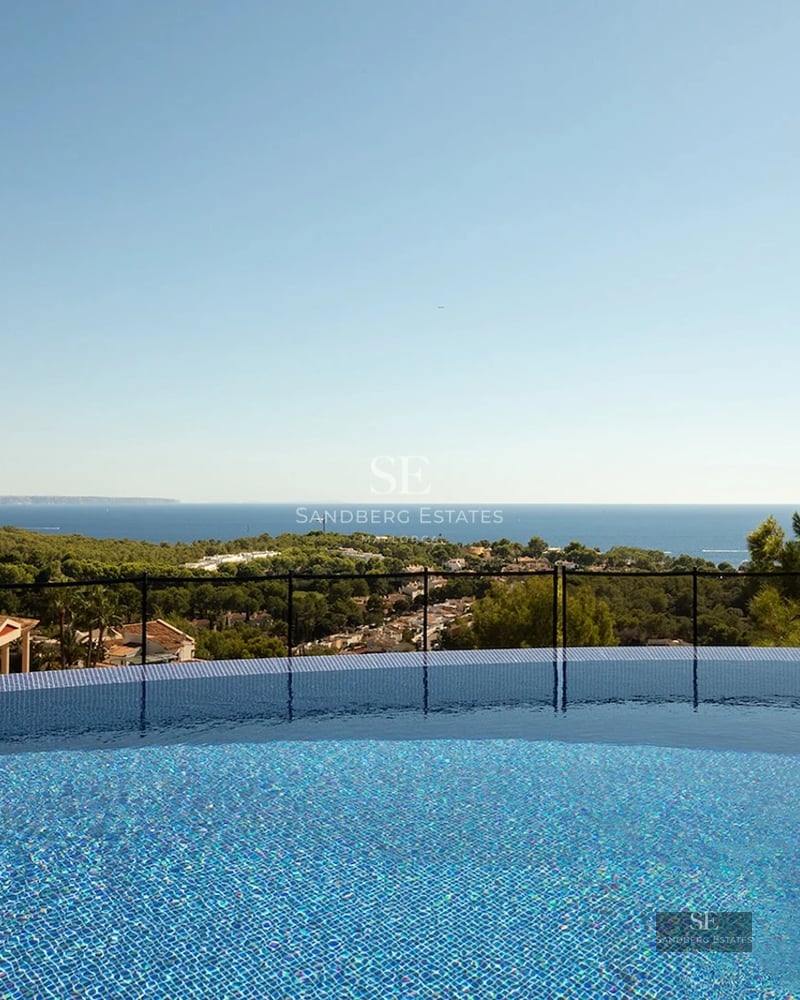 An infinity pool with blue tiles overlooking a lush green forest and the Mediterranean sea under a clear blue sky.